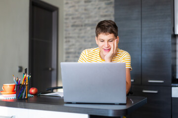 Cute young teenager in yellow shirt sitting behind desk in kitchen next to laptop and study. Serious boy makes homework, listening lesson. Home, distance education, self study by kids.