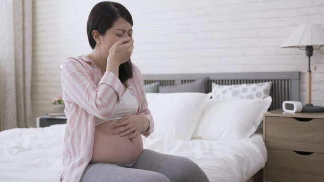 asian pregnant female with her hand on the stomach and mouth is feeling nausea and almost throwing up in the morning at home.