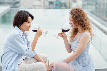 Obraz na płótnie Canvas Beautiful young girlfriends drinking red wine and looking at each other when sitting on rooftop
