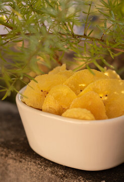Chips de banana frita dentro de un recipiente bajo la sombra de las plantas