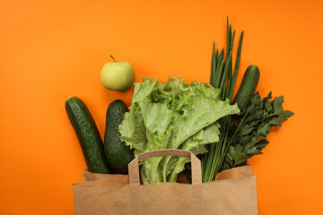 Green vegetables in paper shopping bag on the orange background. Healthy food, full of vitamins. Cucumber, avocado, apple, iceberg salad, parsley and green onion in the products bag. Zero waste. 