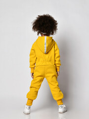 Curly dark-skinned kid girl in warm blue sports jumpsuit with hood and sneakers stands back to camera over white wall background, rear view. Trendy children fashion, stylish outfit - 394563656