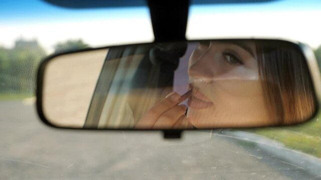 Woman paints lips while looking in the rearview mirror of a car.