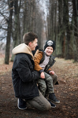 Naklejka na ściany i meble father and son are walking in the park, sitting in the middle of the alley, single father, cold spring, autumn, winter, against the background of large trees, have fun smiling, boy sits on his knees