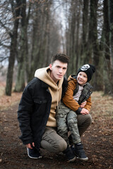 Fototapeta premium father and son are walking in the park, sitting in the middle of the alley, single father, cold spring, autumn, winter, against the background of large trees, have fun smiling, boy sits on his knees