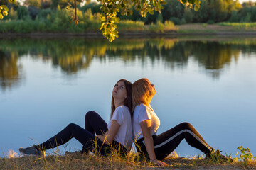 Two athletic young women are resting after training on the river bank. Blonde and the brunette are back to back in nature. Soft focus, blur