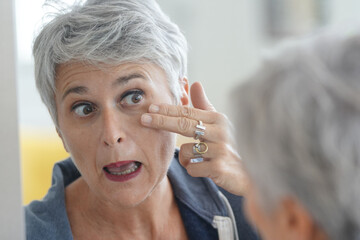 Mature white-haired woman checking eye wrinkles in front of mirror