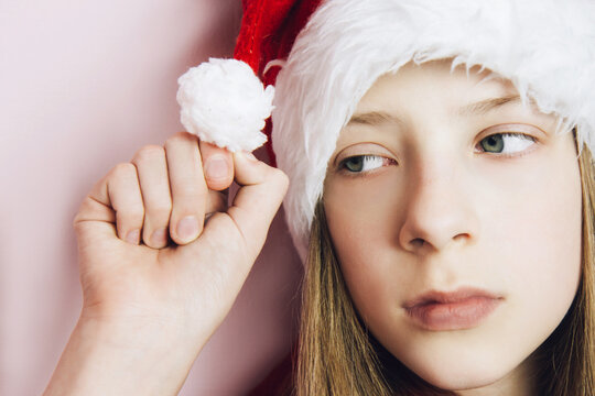 Smiling beautiful girl with long blonde hair in fluffy Santa Claus hat on a pink background. Sad teenage girl holding a bubon on her hat