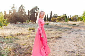 Fototapeta premium Beautiful blonde woman in pink dress walks in the park at sunset