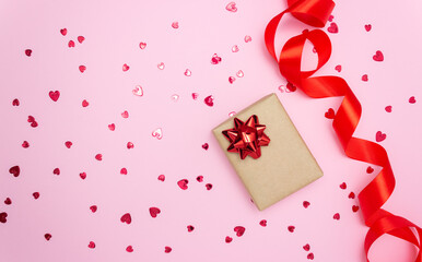Gift box and red satin ribbon on the side on a pink background. Little red hearts are scattered. Free space for text. Gift concept, holidays, love, Valentine's Day.