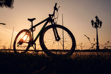 Naklejka premium Silhouette of a bike at sunset. The sun shines through the bicycle wheel with silhouette of big vintage lamppost at sea side park