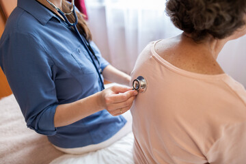 Cropped View of Nurse Putting Stethoscope on Back of Elderly Female Patient in Hospital Room. Back View of Senior Woman Sitting on Bed at Home Having her Breathing Checked by Home Caregiver.