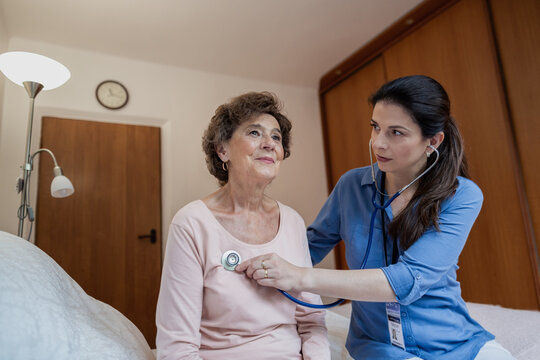 Nurse Listening to Heart of Elderly Female Patient with Stethoscope in Hospital Room. Low Angle View of Senior Woman Sitting on Bed Having her Heart Rate Checked by Home Caregiver.