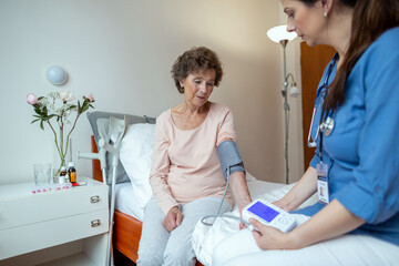 Home Caregiver Checking Blood Pressure of Senior Patient Sitting on Hospital Bed. Home Care Nurse Taking Blood Pressure of Senior Woman With Digital Blood Pressure Monitor in Bedroom.