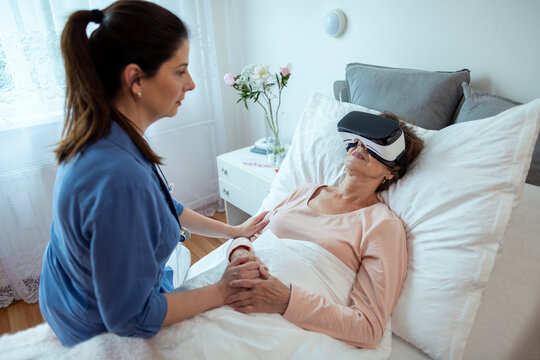Senior Female Patient Relaxing In Hospital Bed with Virtual Reality Headset. Home Nurse Sitting on Bed Looking at Elderly Woman Getting Treatment Via VR Technology.