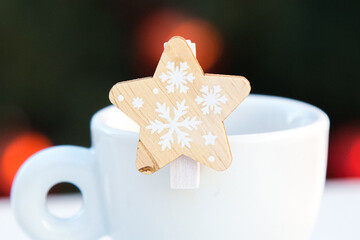 Wooden decorative star with snowflakes closeup on white porcelain coffee cup. Blurred background with red Christmas lights. New year postcard. Congratulations.