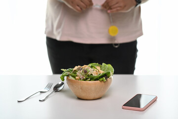 A healthy bowl of salad on white table with a woman measuring her waist size with tape measure.