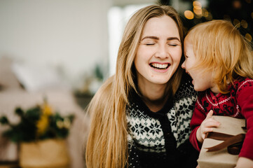 Merry Christmas and Happy Holidays. Mom gives a gift to her daughter near the Christmas tree. Cheerful mother hugging cute baby child girl together at home. Happy New Year 2021.