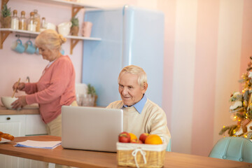 Senior man working on a laptop while his wife cooking