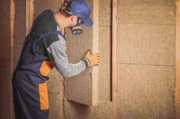 A man in overalls puts mineral sheet insulation between wooden beams. The concept of insulation or sound insulation of walls in frame buildings.