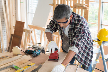 Senior asian old man carpenter using electric wood sanding machine to scrub plank in workshop