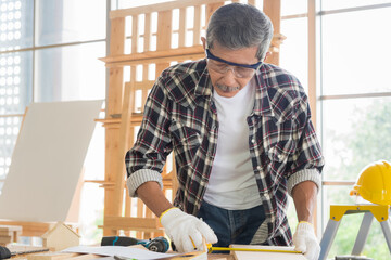 Senior asian old man carpenter using tape measure on wood plank for build diy furniture in workshop