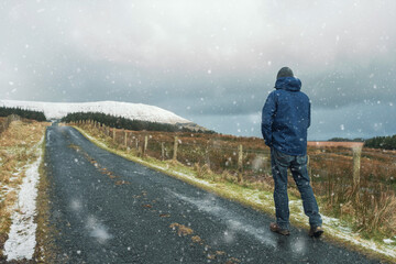 Man in winter clothes walking on a narrow small road to a mountain during snow fall. Winter season concept. Sligo, Ireland.
