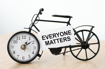 Card with text everyone matters. On the table there is a clock in the shape of a bicycle with a...