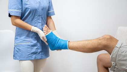 Female doctor in a blue medical gown checking broken leg on male patient.