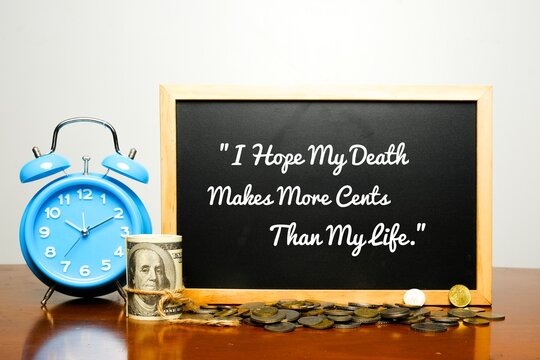 A picture of written word "I Hope My Death Makes More Cents Than My Life" on blackboard with alarm clock, fake money and coins.