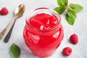 Homemade raspberry jam in a glass jar and fresh raspberries with mint on a grey concrete background. Copy space