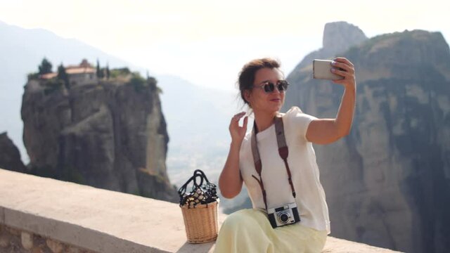 Young woman taking selfies of Meteora rock mountains near Kalambaka, Greece. Large hat, white dress and yellow skirt.