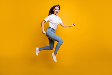 Full length body size photo of young girl jumping high wearing jeans t-shirt sneakers isolated on bright yellow color background