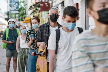 Middle aged man holding longboard, wearing mask waiting, standing in line, respecting social distancing to collect his takeaway order from the pickup point during lockdown