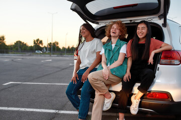 Three young commonly dressed women of different nationalities sitting in an opened car trunk outside on a parking site smiling and laughing
