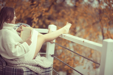 Naklejka premium girl in a knitted sweater reads a book on the veranda in the park, seasonal romance concept