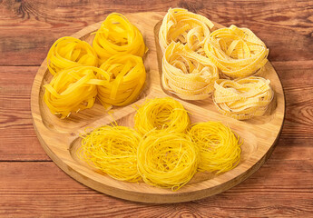 Different uncooked pasta nests on wooden dish on rustic table