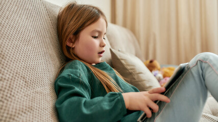 A small girl playing games on her tablet while sitting on a sofa