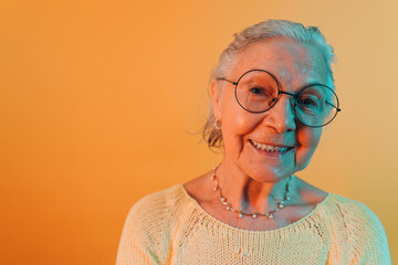 Happy positive old woman in big round eyeglasses smiles with joy looking into camera. Close up portrait of elderly white lady isolated over orange background. Happiness in old age concept.