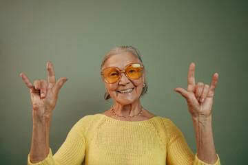 Happy very emotional elderly woman in casual clothes with raised hands looking at camera and smiling. Isolated over green background. Sign of the horns. Music lovers rock fans concept. Orange glasses.