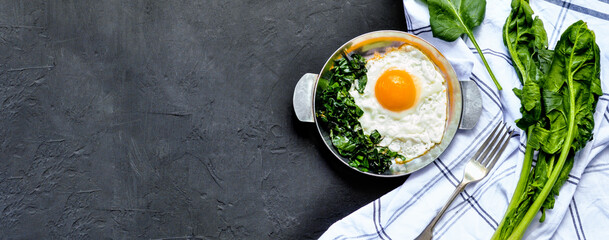 Fried eggs with spinach on iron pan plate. Fried eggs breakfast with white towel and spinach leaves, ingredients on dark black concrete background, Long banner with copy space