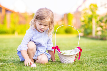 A little cute girl in a blue dress sitting on the lawn collects painted eggs in a wicker basket. Easter egg hunt concept