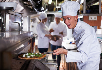 Fototapeta na wymiar Portrait of happy surprised chef getting ready pizza out of professional oven in modern restaurant kitchen