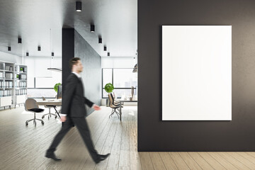 Young businessman walking in workplace with blank banner on wall.