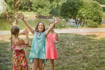Fototapeta premium Beautiful girls play together in the Park and blow soap bubbles. On the background of a large lake.