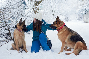 Young woman, wearing blue ski suit, playing with two german shepherds in snow in park. Female master training her dogs in forest with bare trees, squatting down. Winter vacation activities.
