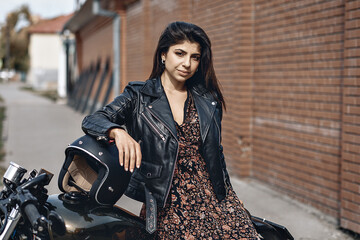 Fototapeta na wymiar Portrait of a cute adorable brunette woman posing on her powerful black motorcycle leaning against a black helmet in a dress and leather jacket. Drive concept