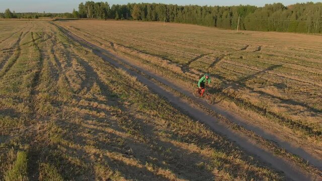 Man in green helmet extreme fast rides make bunny hop jump on orange mtb bike electro mountain bicycle e-bike along rural dirty road dust from wheels past field on sunny autumn evening aerial view.