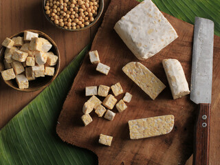 Top View Cut Slicing Raw Tempeh on Wooden Chopping Board, Above Rustic Brown Table. With Banana Leaf, Knife, and Soy Bean