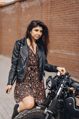 Fototapeta na wymiar Portrait of a sexy force and a bright woman in a black leather jacket and dress posing next to a black retro motorcycle with her knee on the wheel and looking into the distance.